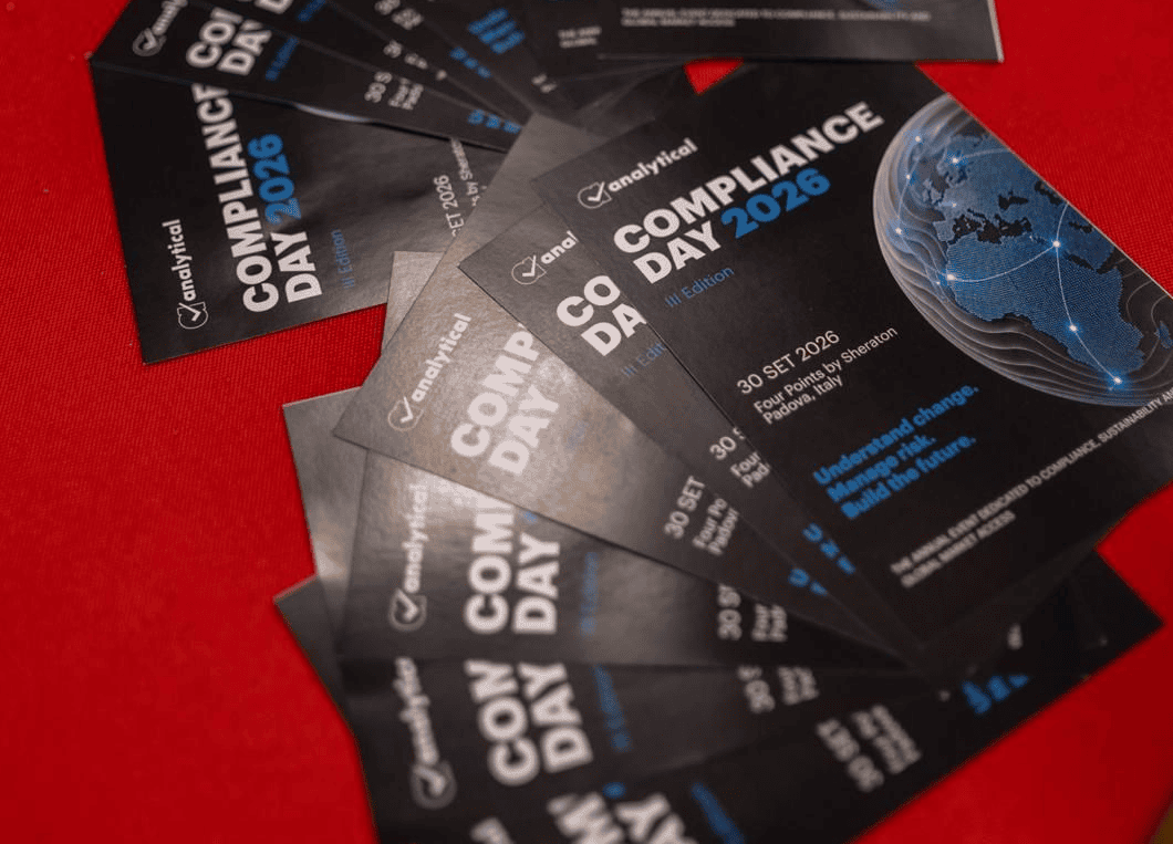 A fan of black and blue event flyers for 'Compliance Day 2026' laid on a red table, with out-of-focus badges and merchandise in the background.
