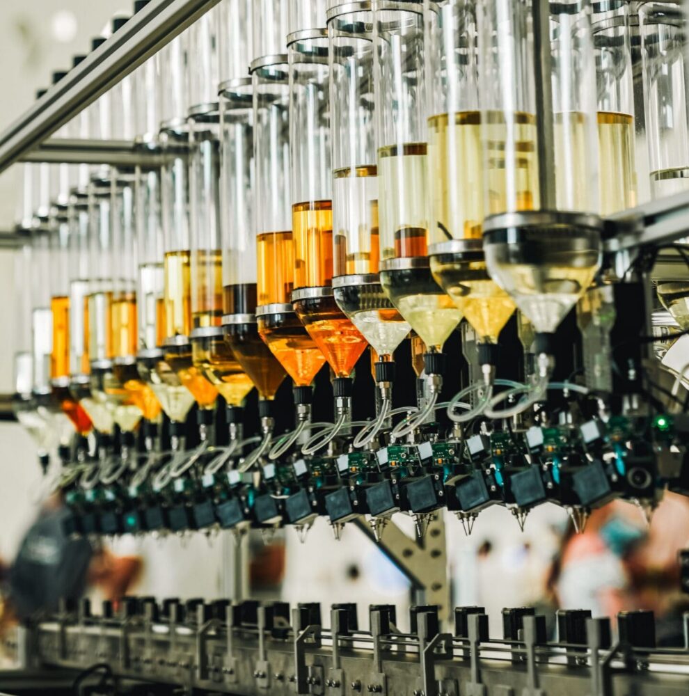 A row of clear glass tubes filled with yellow and orange liquids, part of an automated dispensing machine with electronic components and nozzles, against a blurred background with decorative patterns.
